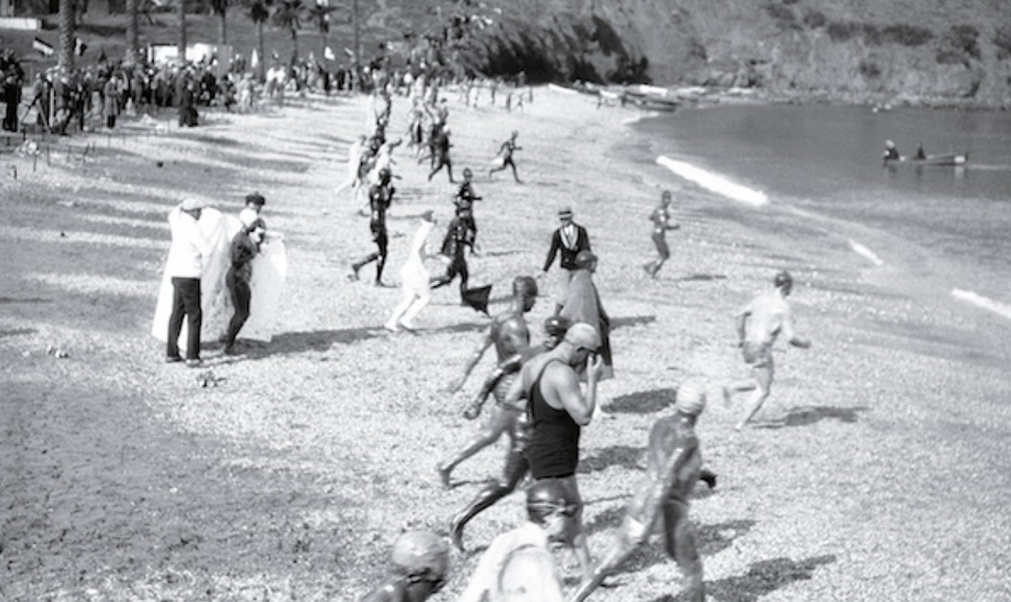 A black-and-white photo of the start of an early 20th-century ocean swimming race. Dozens of swimmers, many covered in grease and wearing minimal or no swimwear, rush across a pebbled beach toward the water. In the foreground, two men hold up a white sheet to shield a nude female swimmer from public view. A crowd of spectators and officials lines the beach under palm trees in the background. A black-and-white photo of the start of an early 20th-century ocean swimming race. Dozens of swimmers, many covered in grease and wearing minimal or no swimwear, rush across a pebbled beach toward the water. In the foreground, two men hold up a white sheet to shield a nude female swimmer from public view. A crowd of spectators and officials lines the beach under palm trees in the background.