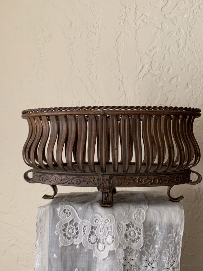 May include: A brown metal oval-shaped basket with a decorative base and three legs. The basket has a woven top rim and vertical bars that form the sides of the basket.