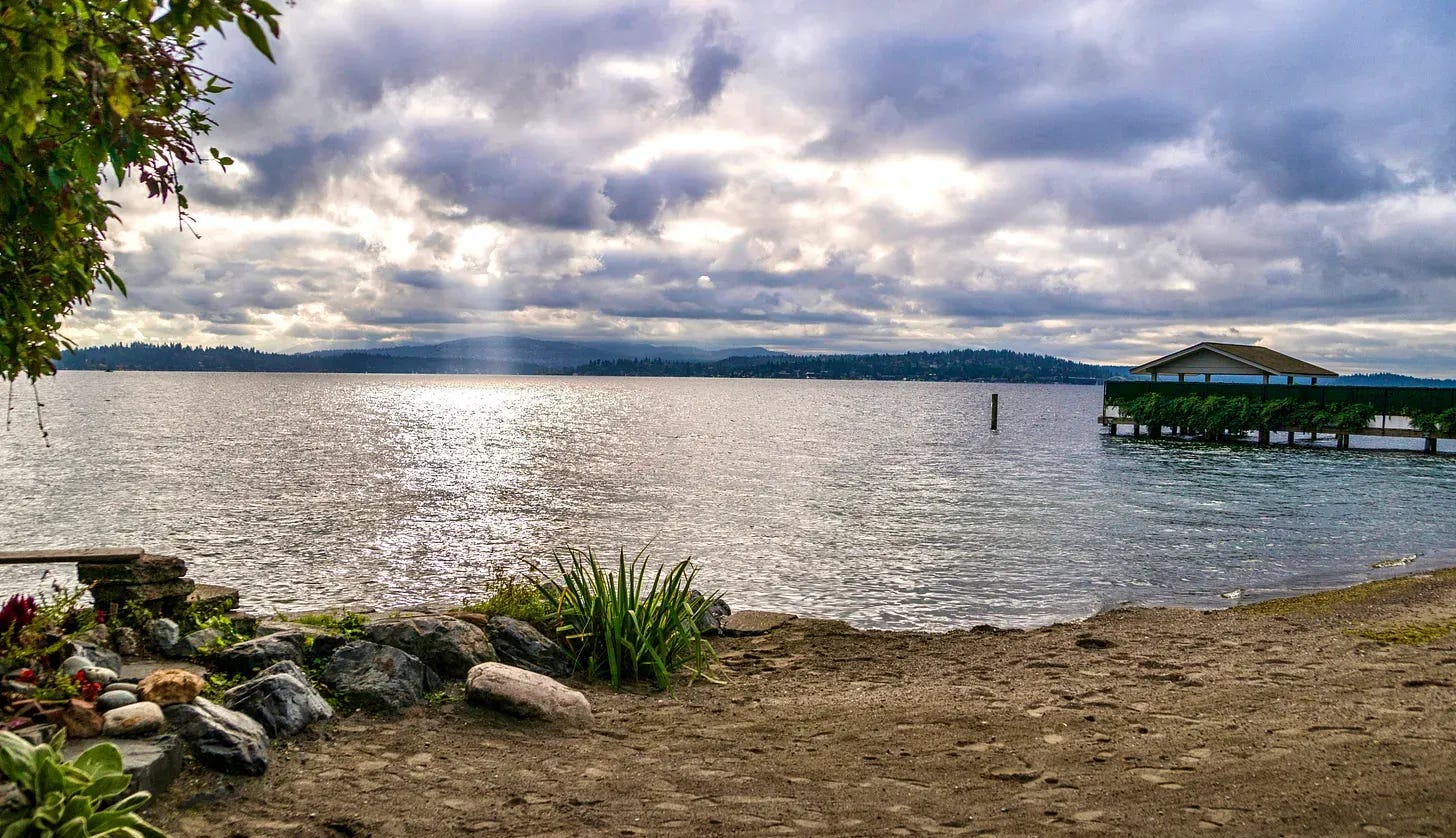 A quiet shoreline at Denny Blaine Park with sandy beach in the foreground, Lake Washington stretching into the distance under dramatic cloud cover, and a small covered dock along the water’s edge.