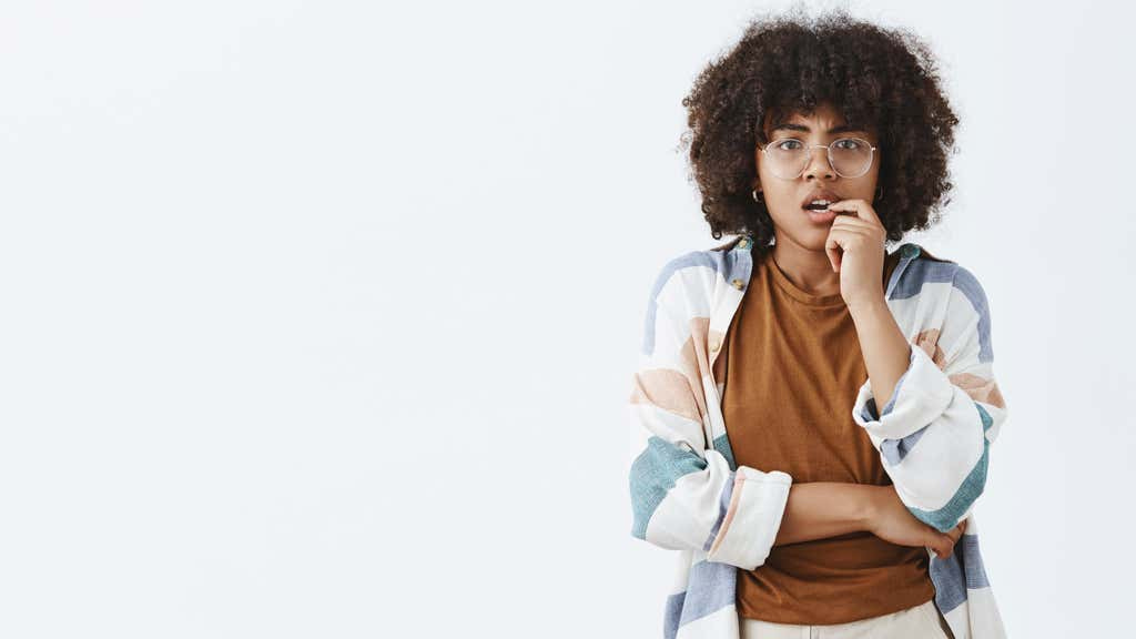 A young black person is standing against a white background, biting their finger and looking anxious