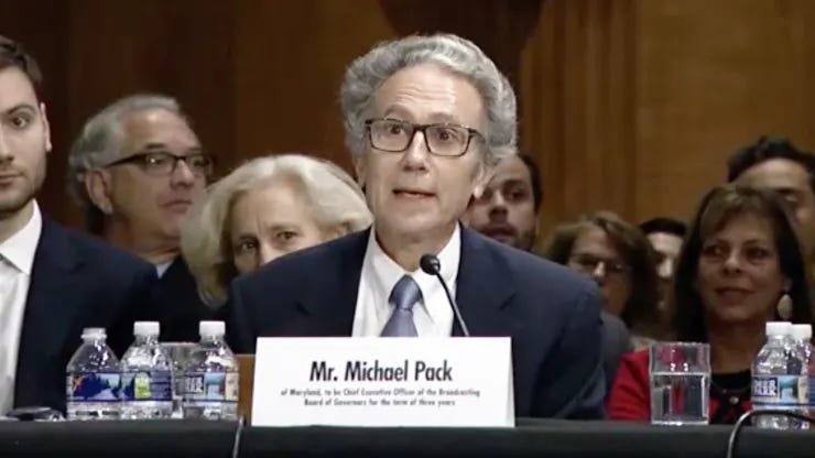 Man with gray-and-white hear who wears glasses speaks before a microphone. Man with gray-and-white hear who wears glasses speaks before a microphone.