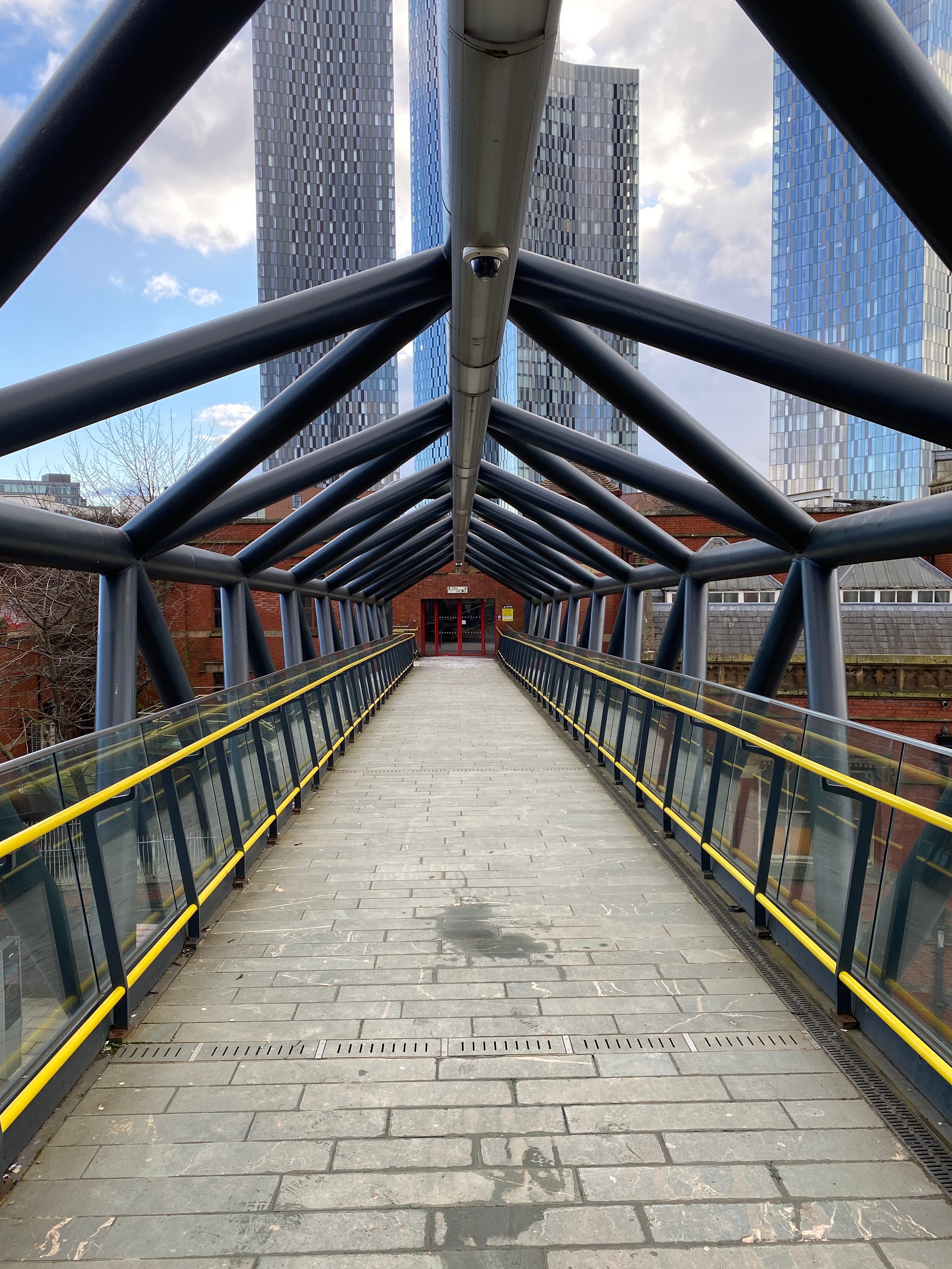 A covered pedestrian bridge with a glass and steel frame leading toward a red doorway. Four tall modern towers rise behind it, their surfaces reflecting a cloudy sky. The walkway is empty, and the lines of the structure draw the eye forward.