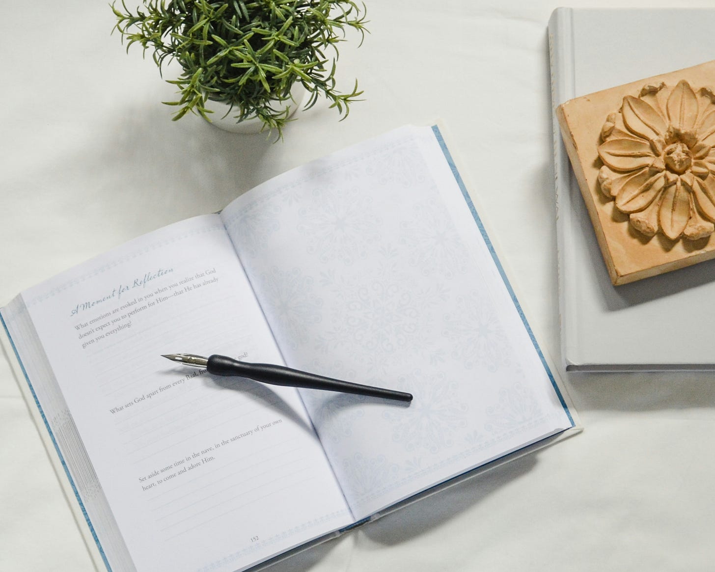 Open journal with a black pen on top, resting on a white surface. To the side, a potted plant and a decorated book create a calm, reflective atmosphere.
