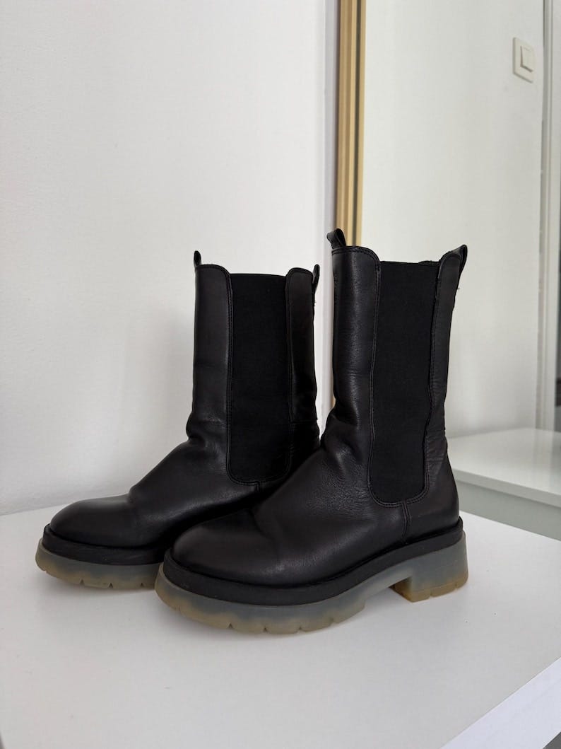 May include: A pair of black Chelsea boots with a chunky, transparent sole. The boots have a smooth leather upper, elastic side panels, and a rounded toe. The soles are thick and have a slight platform, providing a modern look. The boots are displayed on a white surface.