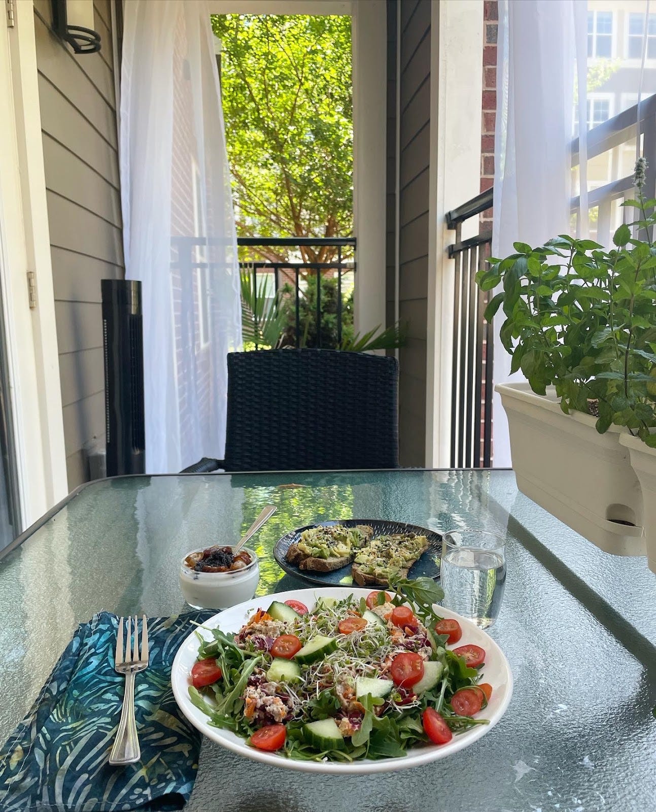 A salad is pictured on a well lit outdoor patio table with a pot of herbs set to the side.