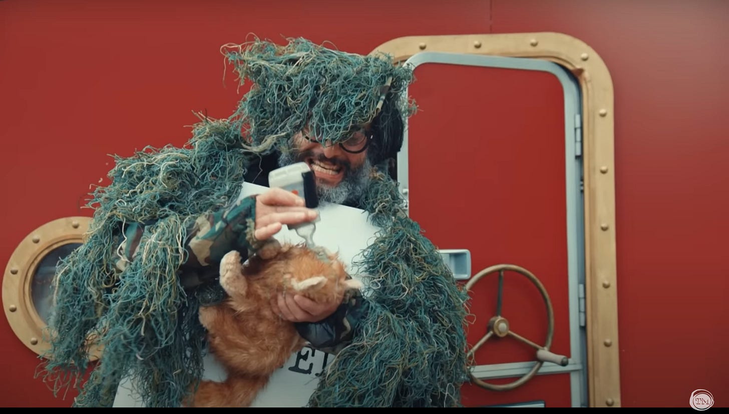 Jason Mantzoukas is dressed in a wild-haired costume as he bottle feeds Patatas the stuffed cat.
