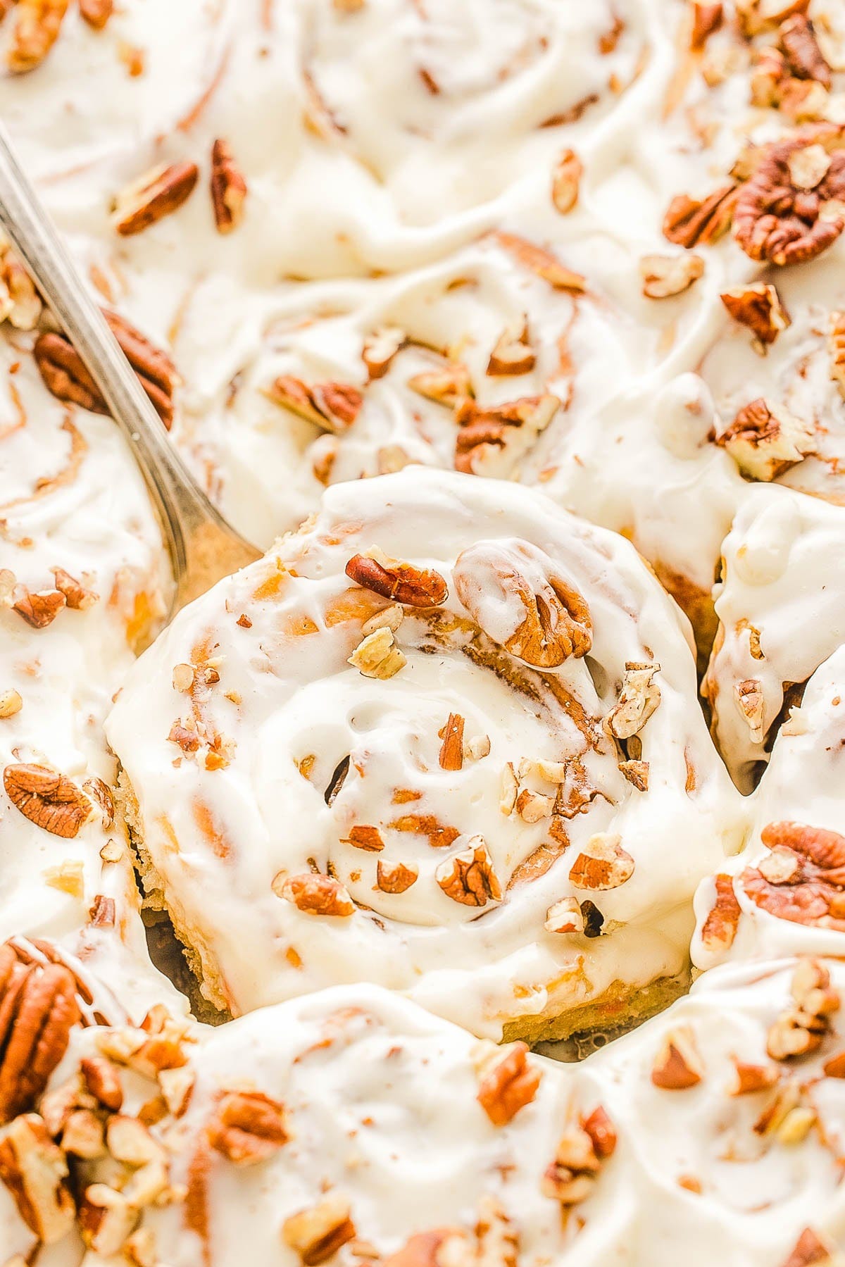 Close-up of frosted cinnamon rolls topped with chopped pecans, with a serving spoon lifting one.