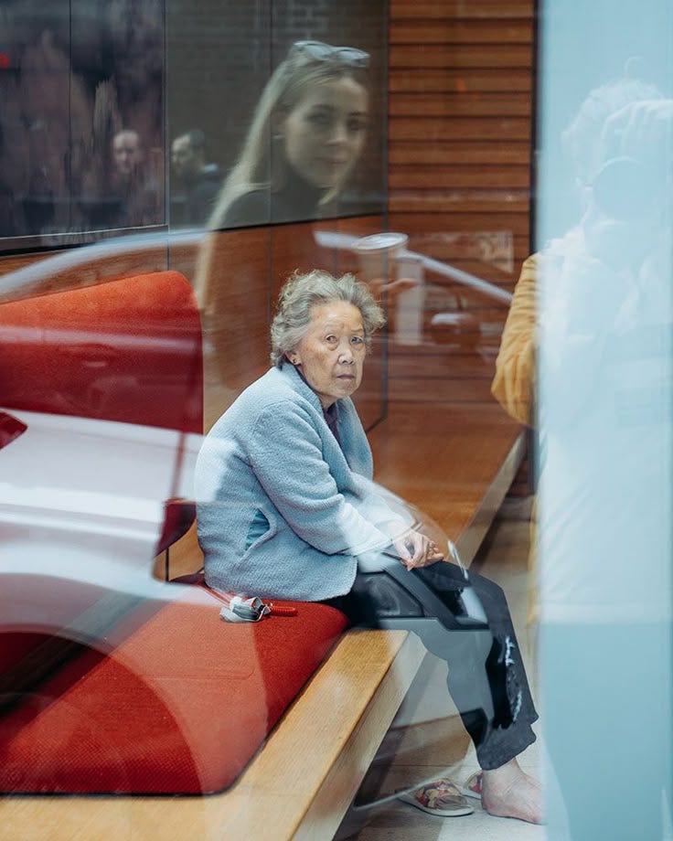 This may contain: an older woman sitting on a bench in front of a glass window looking at her reflection