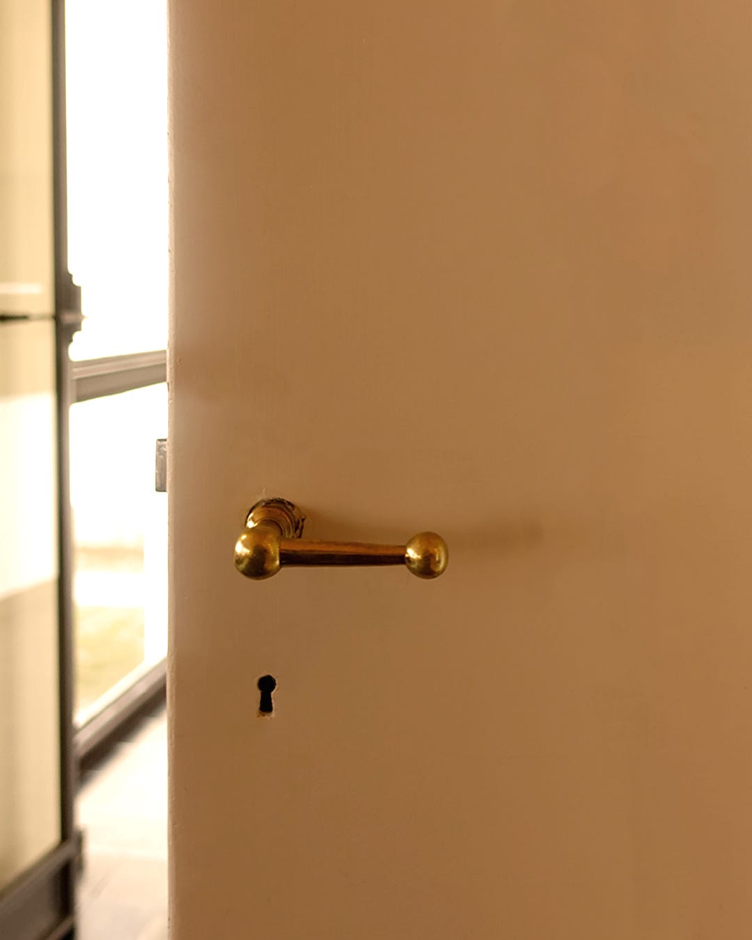 Close-up of an antique brass door handle and keyhole on a white door, bathed in soft golden light from an adjacent bright window
