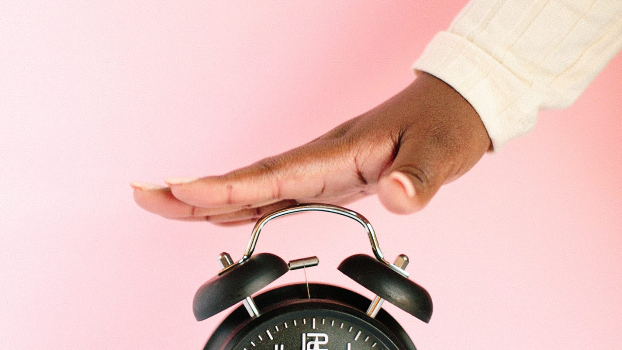 Black hand hovering over a ringing alarm clock against a pink background