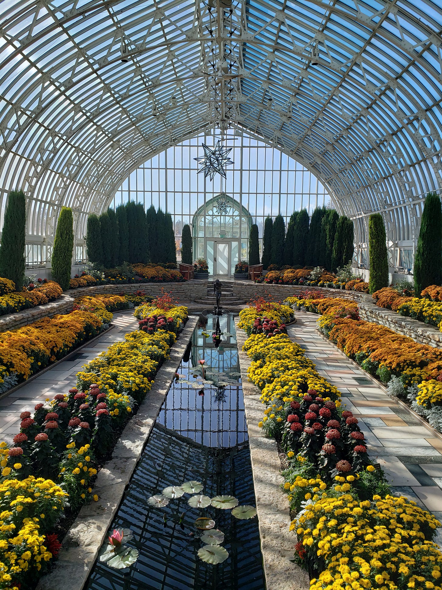 Looking into the Sunken Gardens from ground level with various yellow, orange, and red flowers around the main water feature and statue.