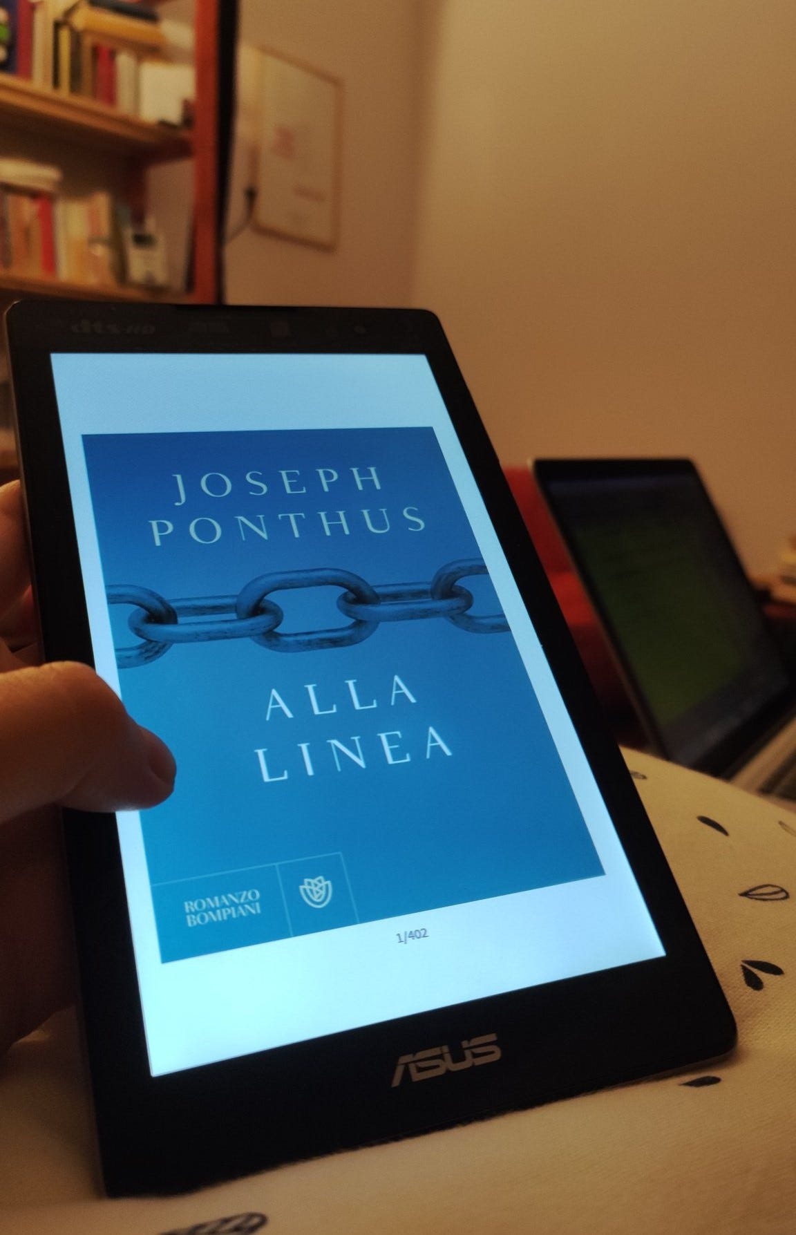 In primo piano una mano tiene un tablet. Sul tablet si vede la copertina di un libro, blu: è alla linea di joseph ponthus. Dietro si intravede un'altra persona con il computer. Sullo sfondo c'è una libreria in legno piena di libri