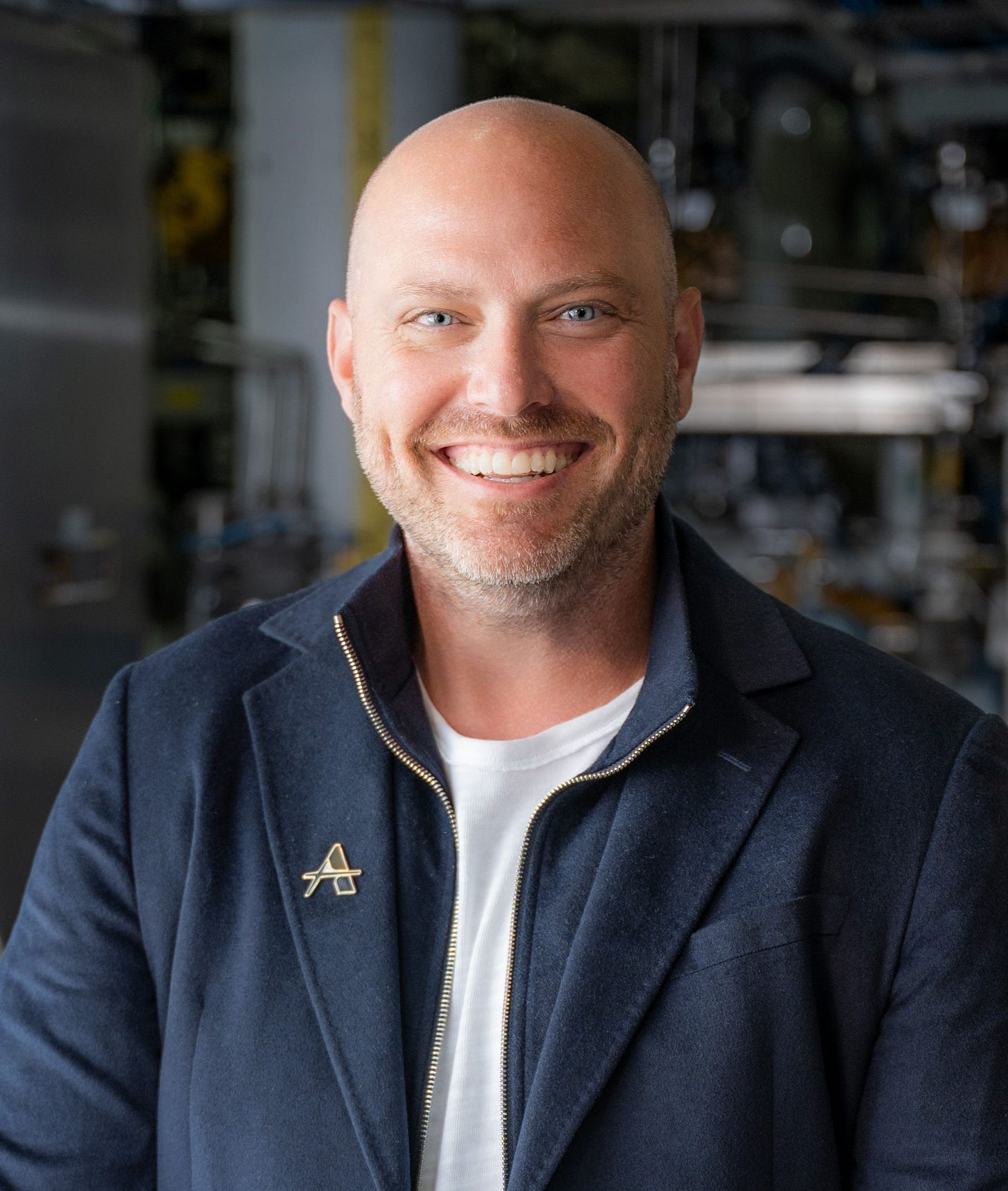 Chris Newton, Chief Engineer at Applied Atomics, in a navy jacket with an Applied Atomics lapel pin, standing in front of piping and equipment at the IST facility.