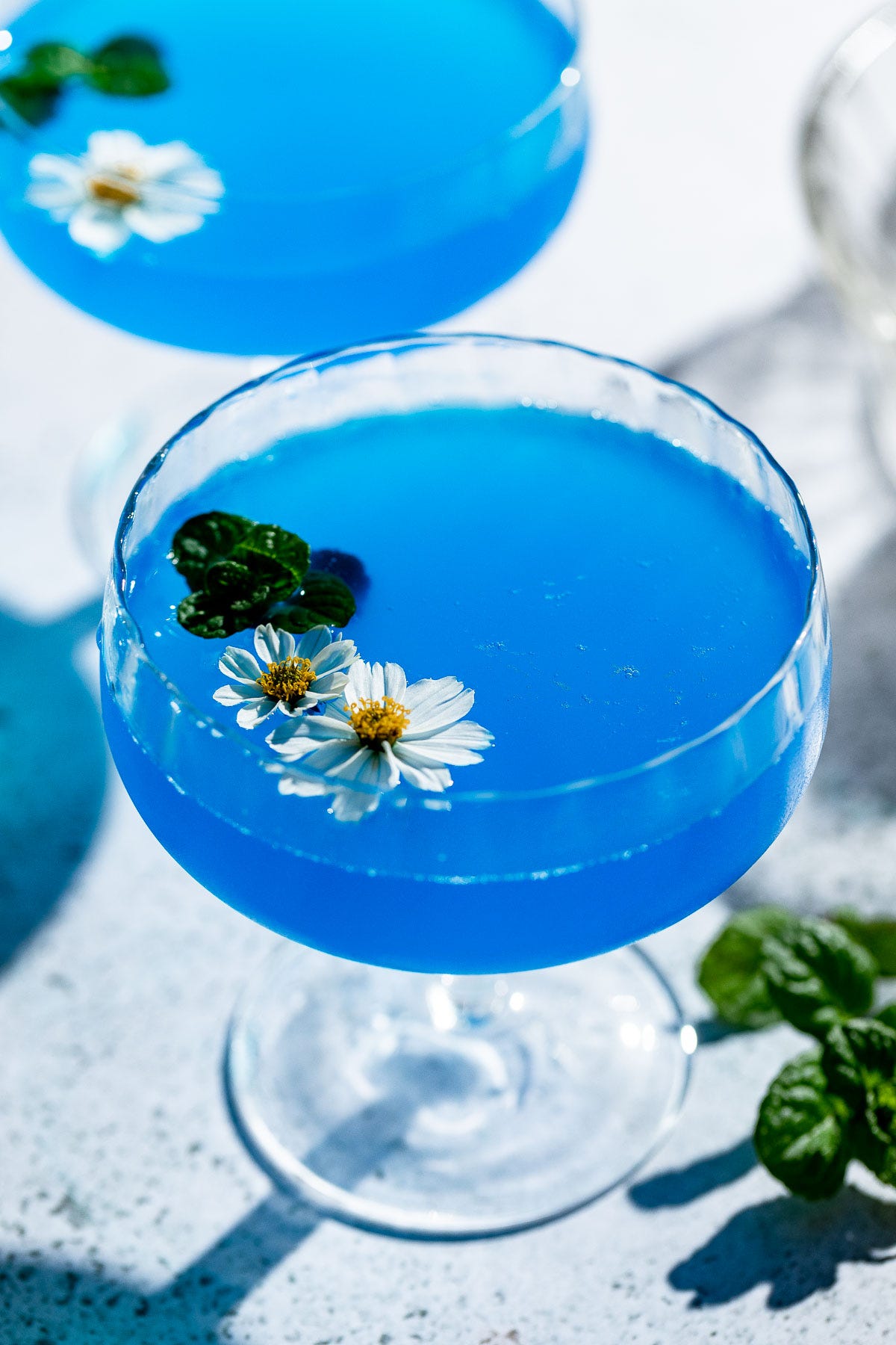 Two blue lagoon cocktails served in coupe glasses with fresh flowers.