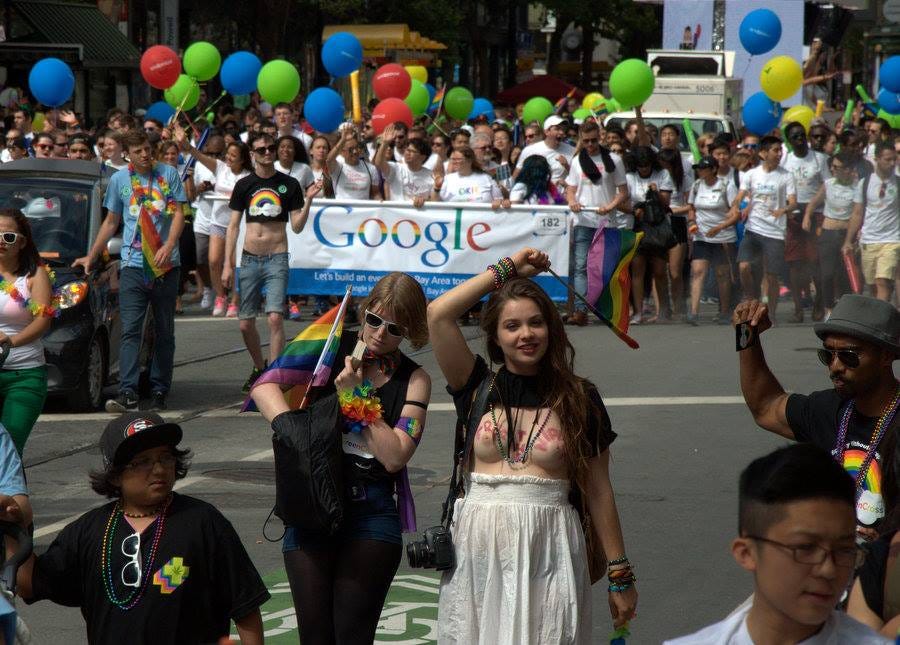San Francisco'daki bir onur yürüyüşünden bir foto. Önde iki kadın, ellerinde bayraklar, birinin göğüsleri açık, yüzünde bir gülümseme. Arkada Google şirketinde çalışanların korteji geliyor, ellerinde balonlar. San Francisco'daki bir onur yürüyüşünden bir foto. Önde iki kadın, ellerinde bayraklar, birinin göğüsleri açık, yüzünde bir gülümseme. Arkada Google şirketinde çalışanların korteji geliyor, ellerinde balonlar.
