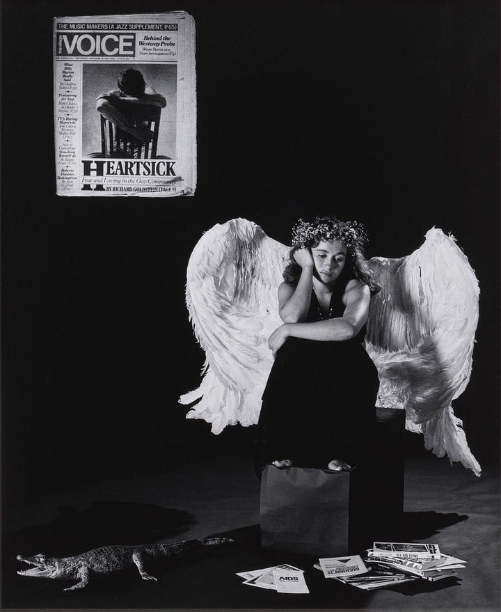 1) A woman with large white angel wings and a floral crown sits pensively amid scattered newspapers and a small faux alligator. A newspaper headline about an AIDS victim is featured on the wall behind the angel. The scene conveys a somber, reflective tone. The angel is holding her head up with her hands. The photo is in black and white, and the angel is barefoot, sitting on a box. 2) A woman wearing large, white angel wings, and a flower crown, sits barefoot pensively on a box. There's a newspaper with David Wojnarowicz on the wall behind her and a small faux alligator on the ground in front of her along with several AIDS and safe sex pamphlets, creating a surreal scene. The photo is in black and white.