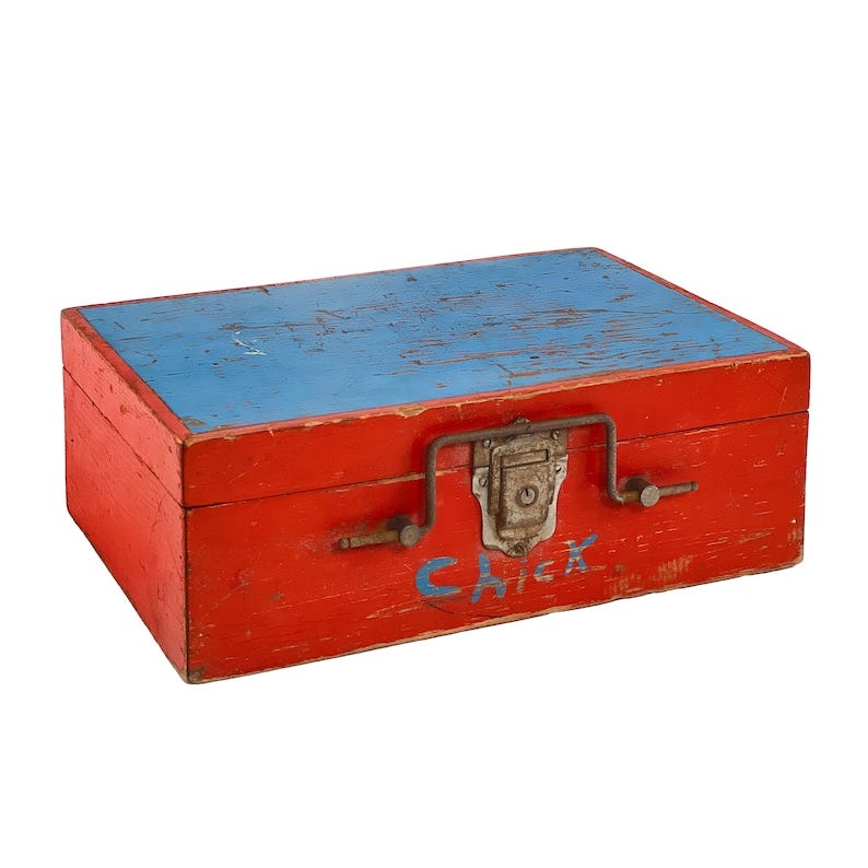 May include: A red wooden box with a blue lid. The lid has a worn, distressed look. The box has a metal latch and a handle. The box is labeled 'Crick' in blue paint.