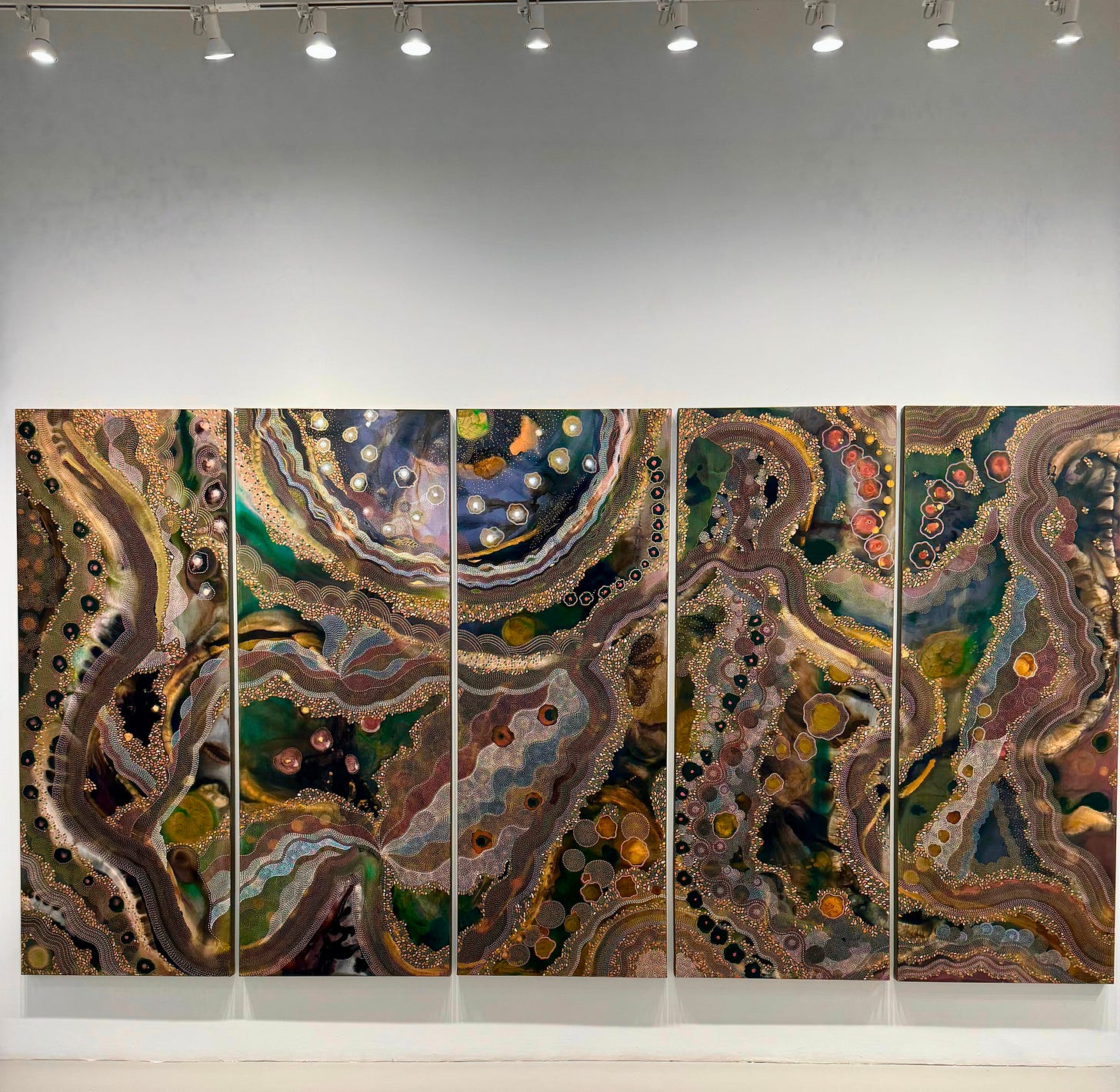 Five-panel abstract work by Trishla Jain installed on a white gallery wall. The composition is filled with layered organic forms, fine dotted patterns, and swirling bands of green, brown, gold, blue, and rust beneath a row of gallery lights.