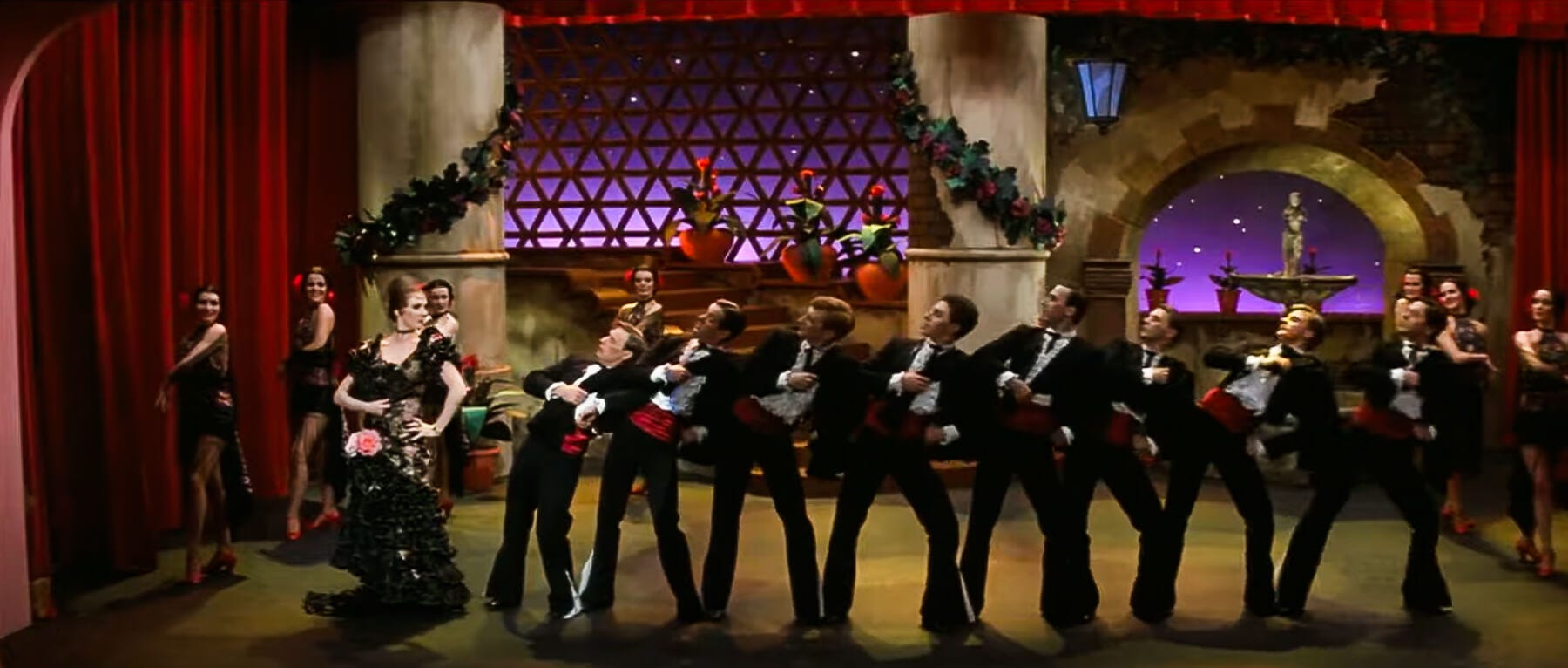 Julie Andrews, dressed as the Shady Dame from Seville, leads a line of tuxedoed backup dancers in a dazzling nightclub performance with rich reds and golds.