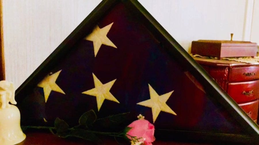 Framed triangular flag with five white stars on a deep red background displayed on a surface next to a small wooden box with a knob, a wooden dresser, a white vase with green leaves and a pink rose, and a dark green leaf in the foreground.