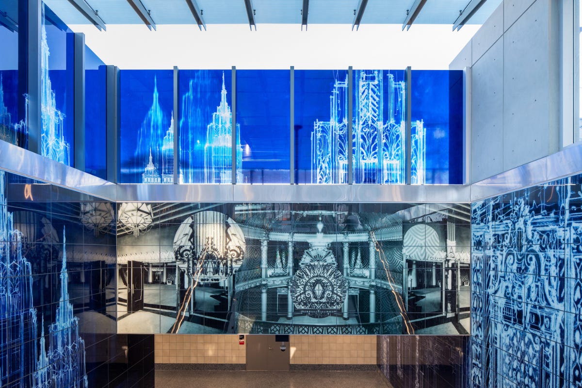 A vivid blue and black station artwork shows chalky white architectural drawings, theater interiors, and layered textile-like patterns across glass and wall panels. A vivid blue and black station artwork shows chalky white architectural drawings, theater interiors, and layered textile-like patterns across glass and wall panels.
