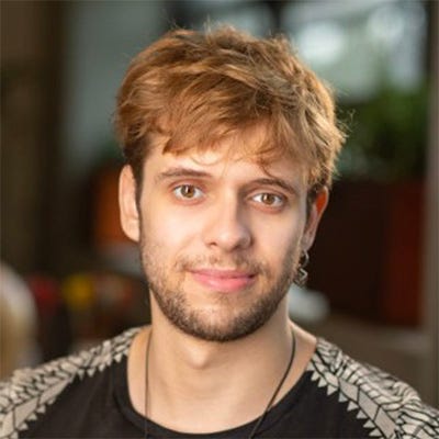 Homem branco com cabelo claro e barba em frente a um fundo destacado utilizando camiseta preta com detalhes brancos nas mangas.