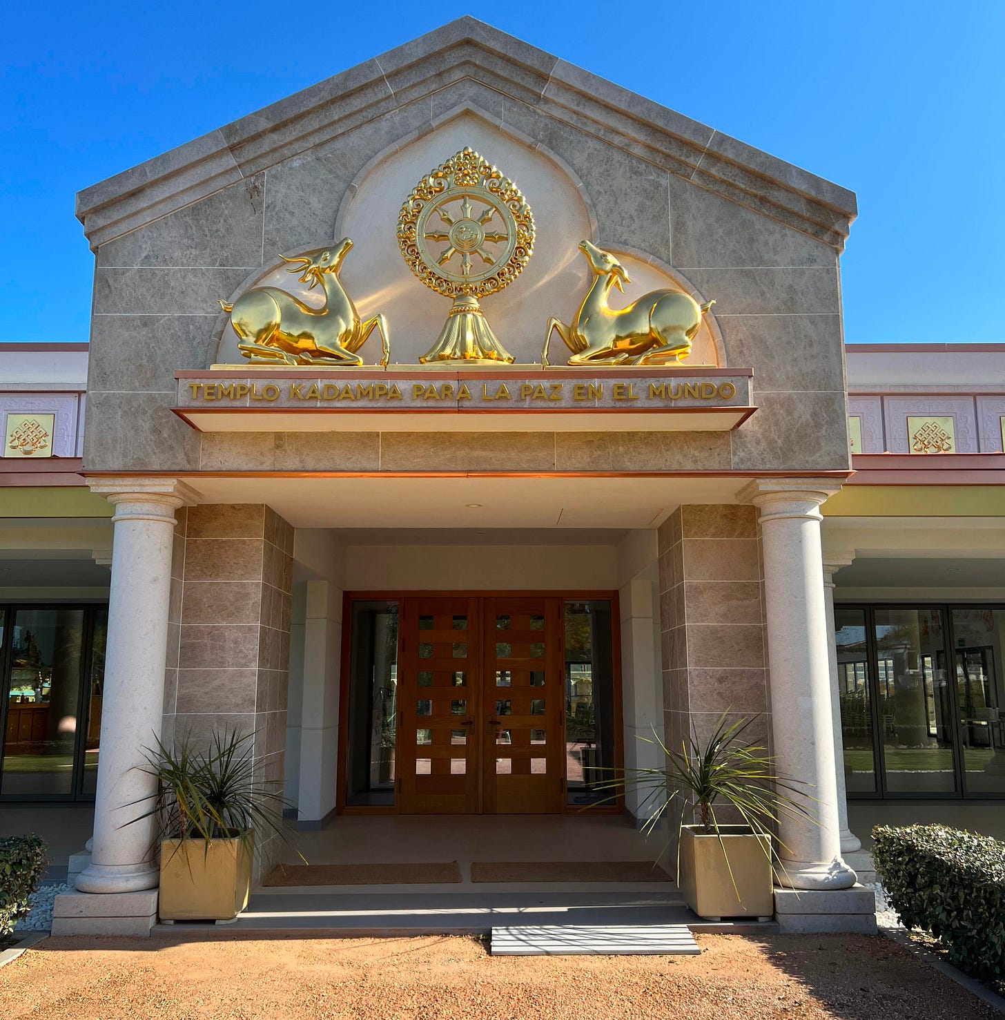 Gold ornamental deer and Dharma wheel above the entrance of Templo Kadampa Para La Paz En El Mundo, with columns and wooden doors below a clear blue sky. Gold ornamental deer and Dharma wheel above the entrance of Templo Kadampa Para La Paz En El Mundo, with columns and wooden doors below a clear blue sky.