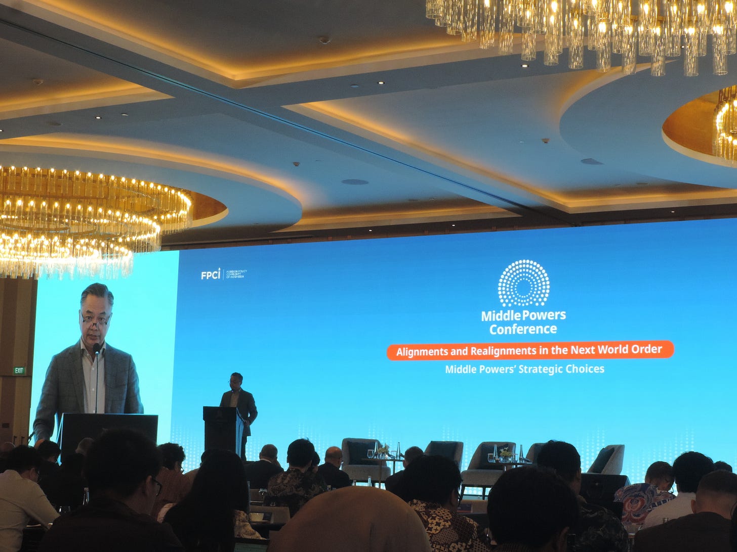 A medium-long shot captures a formal setting during the Middle Powers Conference. Two men are present on a lit, elevated stage in an opulent conference hall; the man to the left of the image is at a podium speaking to an indistinct audience of approximately twenty people at various tables, and the man to the right stands next to another podium in the background. Beyond them is a massive, lit screen with a gradient of blue and white. Text on the screen reads, "MIDDLE POWERS CONFERENCE Alignments and Realignments in the Next World Order Middle Powers' Strategic Choices." The bottom row of text on the screen is in white text set inside an orange rectangle with rounded edges. Three gold, multi-tiered chandeliers are visible at the upper left, center-left, and center-right of the room.