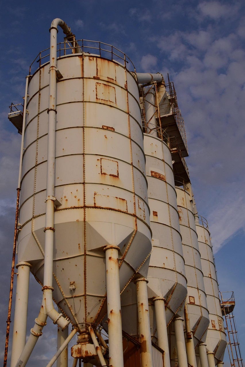 A row of large, weathered industrial silos with rust patches, viewed from below against a partly cloudy sky. A row of large, weathered industrial silos with rust patches, viewed from below against a partly cloudy sky.