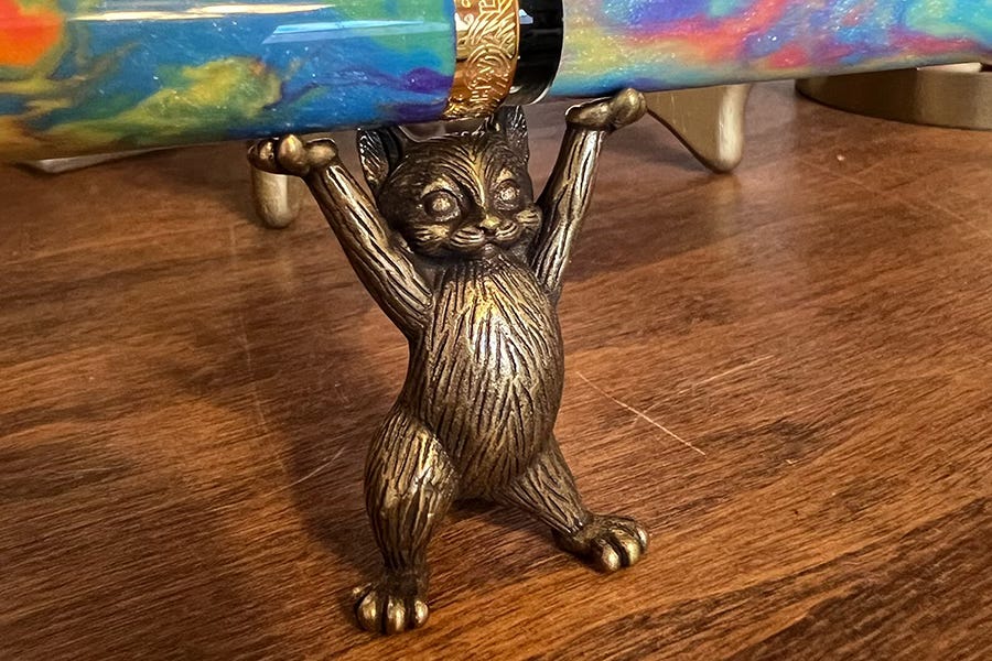 bronze figure of a cat standing on its hind legs, holding it's front paws over its head; it's holding a fountain pen.