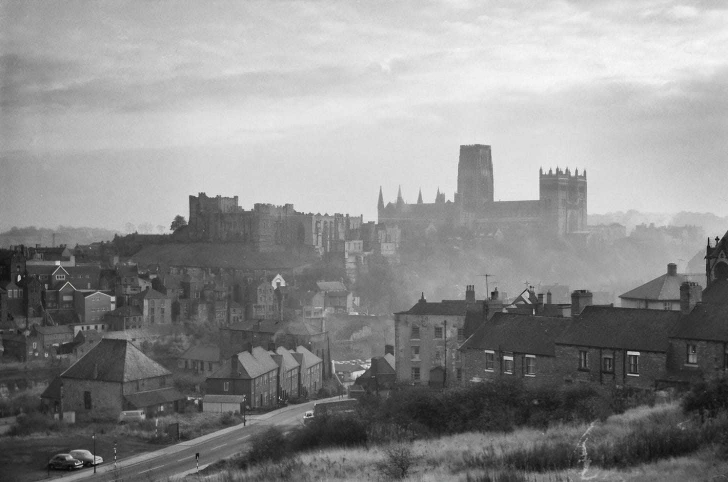 Black and white photograph of a landscape featuring a historic castle and cathedral set against a cloudy sky, with houses in the foreground.