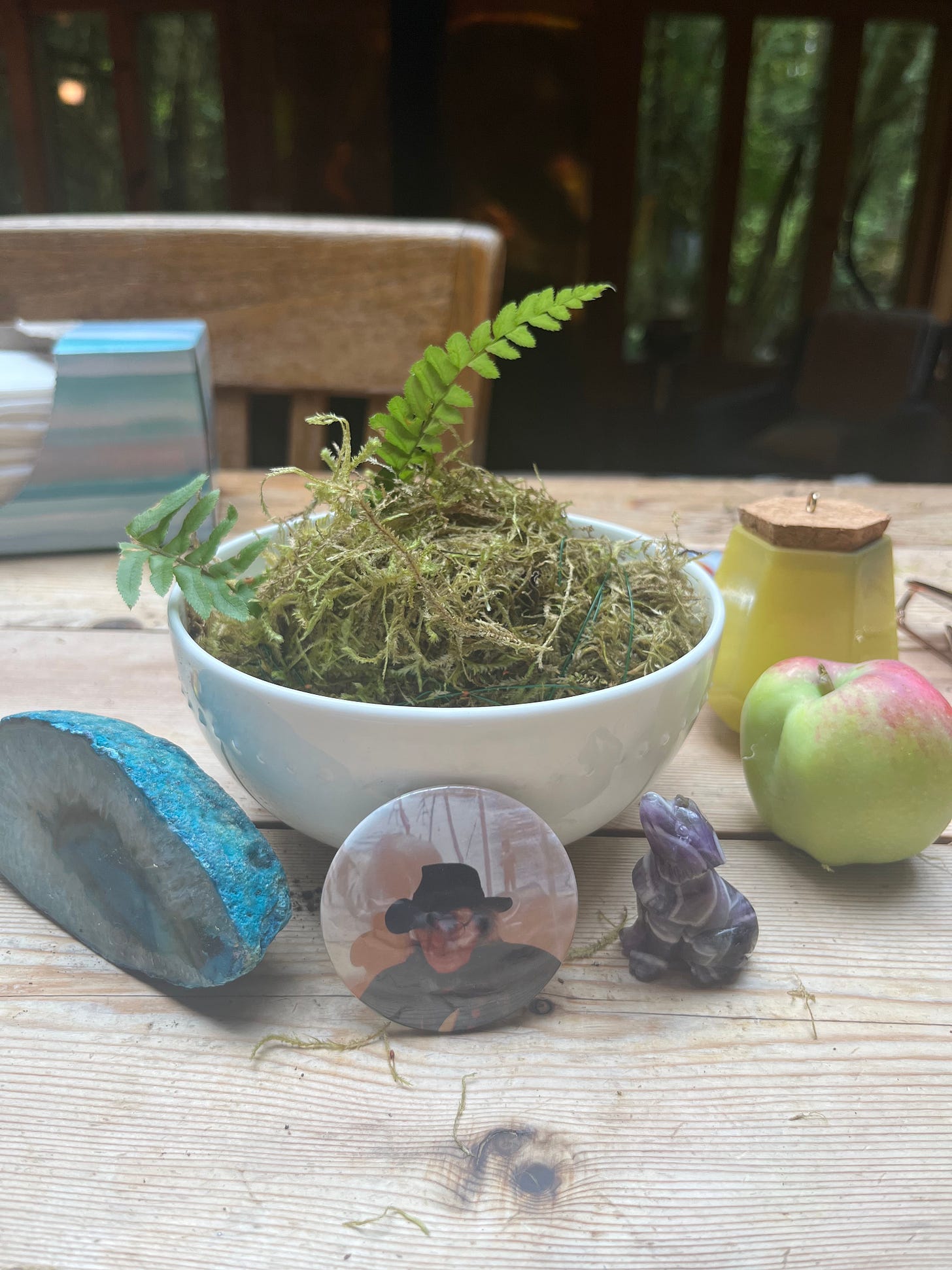 A bowl of moss sits on a table surrounded by small items. Propped up against the bowl of moss is a button pin with a photo of Jack Hirschman. A bowl of moss sits on a table surrounded by small items. Propped up against the bowl of moss is a button pin with a photo of Jack Hirschman.