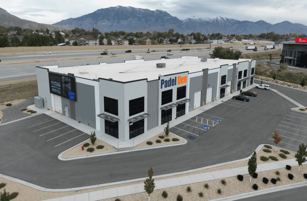 Aerial drone view of Padel Den USA facility in Utah showing modern commercial building with prominent Padel Den signage, blue and orange branding, parking lot with vehicles, highway access, residential neighborhood, and snow-capped mountain backdrop Aerial drone view of Padel Den USA facility in Utah showing modern commercial building with prominent Padel Den signage, blue and orange branding, parking lot with vehicles, highway access, residential neighborhood, and snow-capped mountain backdrop