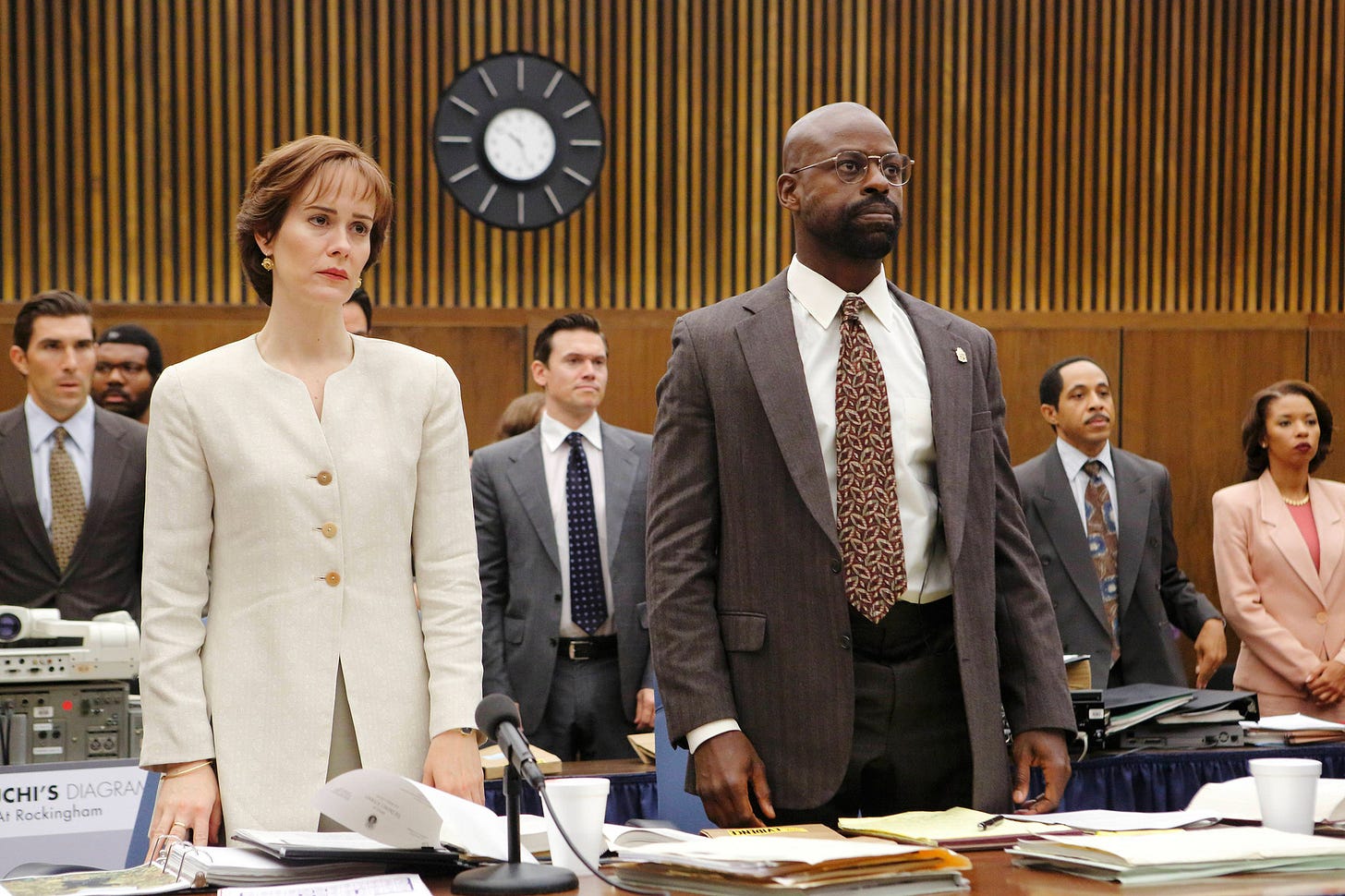 A white woman wearing a beige suit, played by Sarah Paulson, stands next to a black man wearing a grey suit and patterned tie, played by Sterling K. Brown. They're in a courtroom, and other people can be seen behind them.