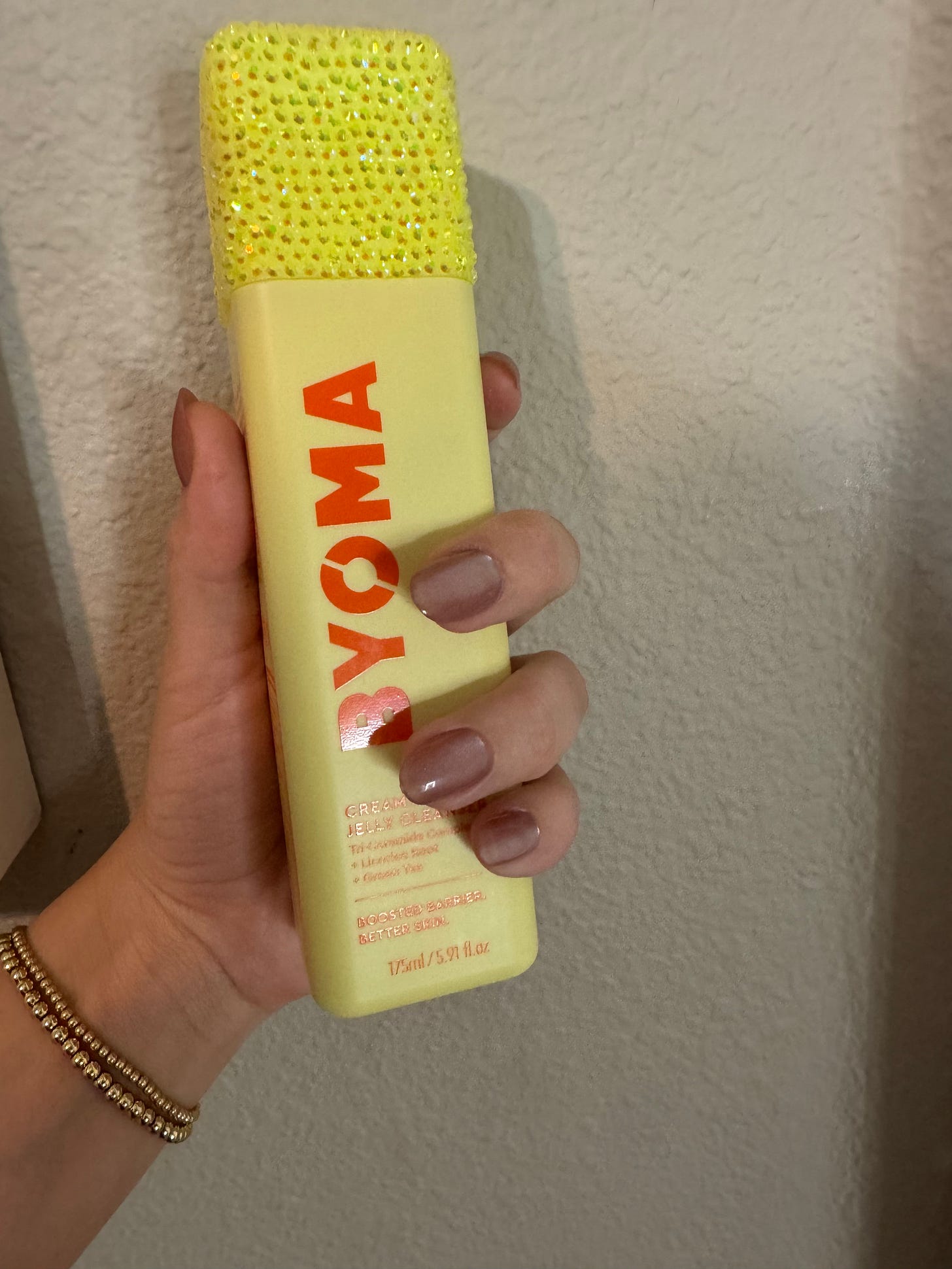 A close-up of a Byoma Creamy Jelly Cleanser bottle customized with a bright yellow bedazzled cap, held by a student with a neutral-toned manicure—a perfect example of Gen A’s ‘Main Character’ product customization.
