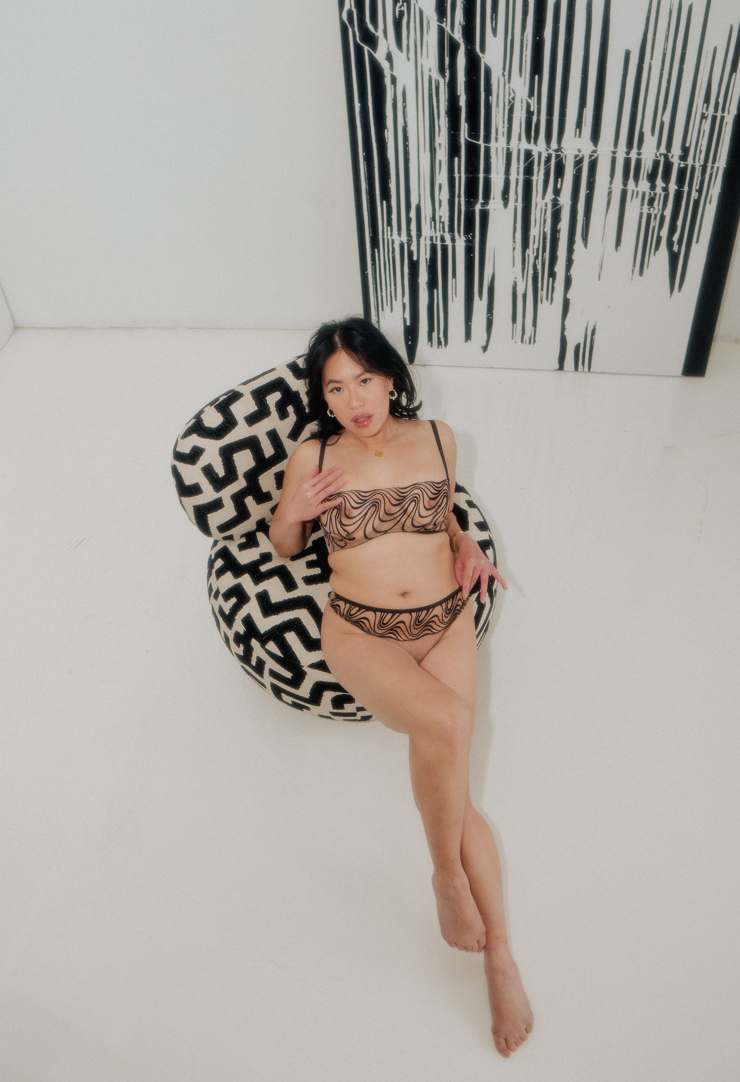 Josie Qu is an asian woman with mid length black hair and full lips looking up at the camera. She is wearing nude mesh lingerie with a black abstract embroidery. She is sitting on a geometric upholestered chair and a black and white abstract painting.