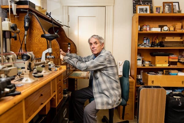 An older man in a gray plaid shirt sits at a desk in a room of wooden furniture covered with tools. An older man in a gray plaid shirt sits at a desk in a room of wooden furniture covered with tools.