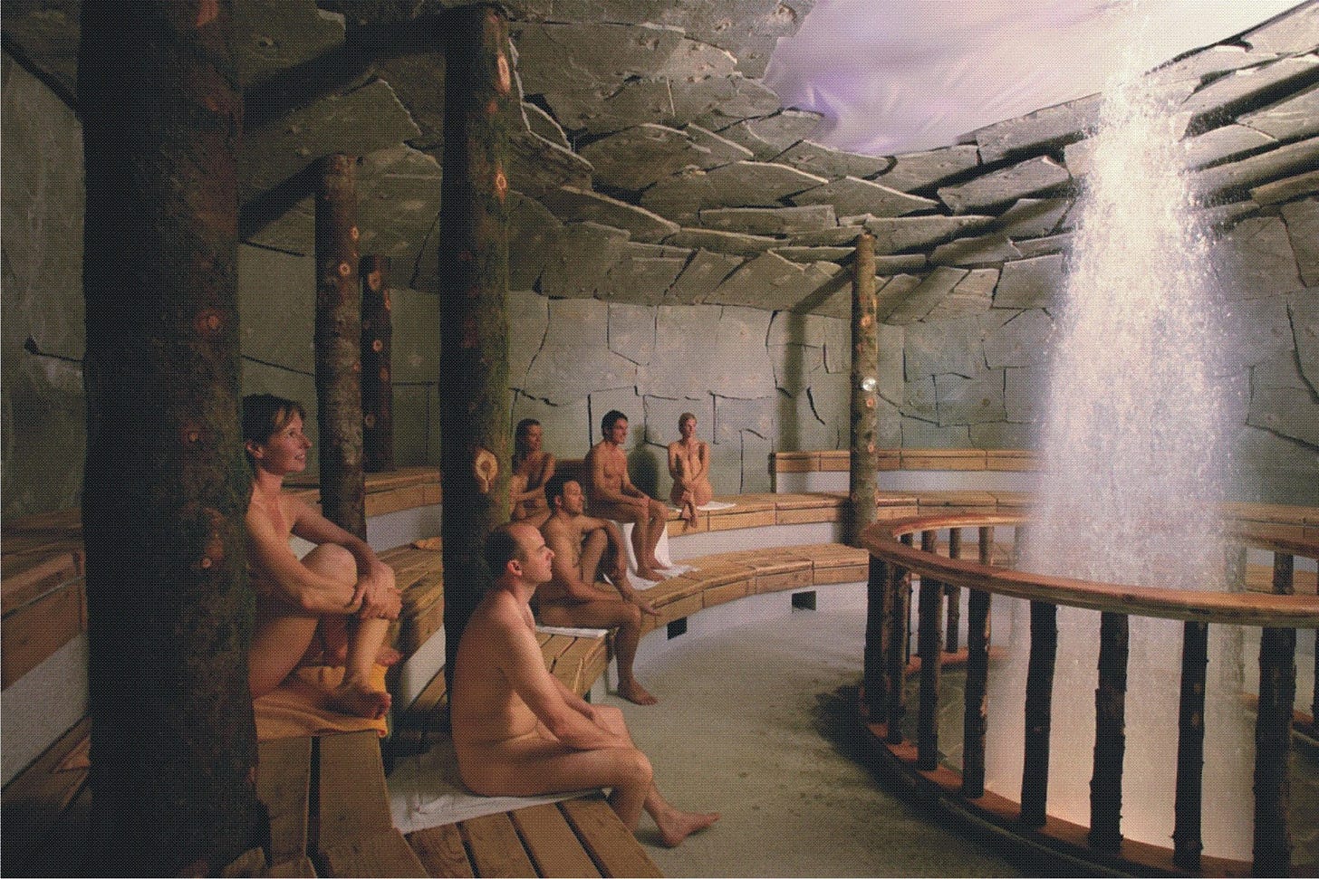 Six nude guests sit on tiered wooden benches inside the Geysirhöhle, a cave-like sauna at Therme Erding. The circular room features rough stone walls, a slate-slab ceiling with purple ambient lighting, and rustic tree-trunk support columns. To the right, behind a bamboo railing, a geyser shoots a column of water upward. The guests sit quietly, facing the geyser feature. Towels are placed on the benches beneath them.