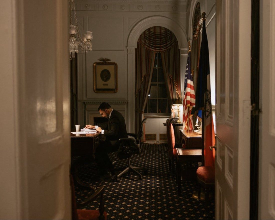 Zohran Mamdani sits alone at a wooden desk in his mayoral office late at night, leaning forward as he reviews papers under a desk lamp. The photo is framed through an open doorway, emphasizing a quiet, private moment. An American flag and a city flag stand to the right, while tall draped windows, chandeliers, and traditional furnishings give the room a formal, historic feel. The lighting is warm and subdued, conveying focus and solitude.