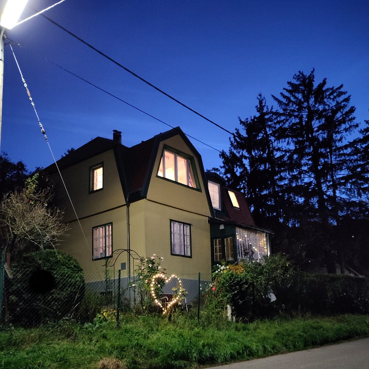 A yellow-painted house is surrounded by dark trees. It is starting to get dark, but there is a street light in the left upper corner and a winter light on the fence, shaped like a heart.