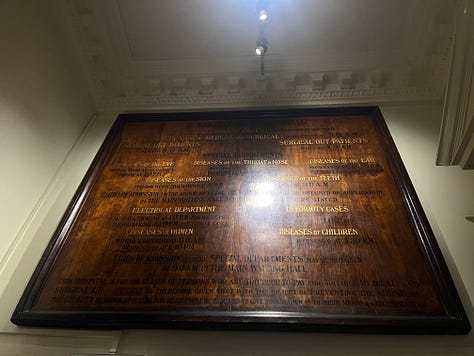 The door to the Barts Museum, a plaque commemorating Holmes and Watson's first meeting, and a large wooden sign advertising medical care.