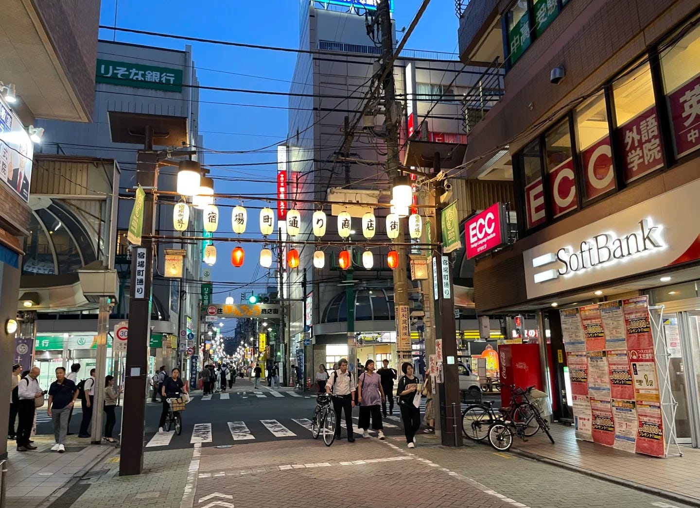 A lantern-strung shopping street in Kita-Senju, Adachi ward, at dusk, with commuters walking home past low-rise shopfronts and parked bicycles. Adachi is among the cheapest wards in this post's rent table, at ¥52,000 (about $325) for a 1K.