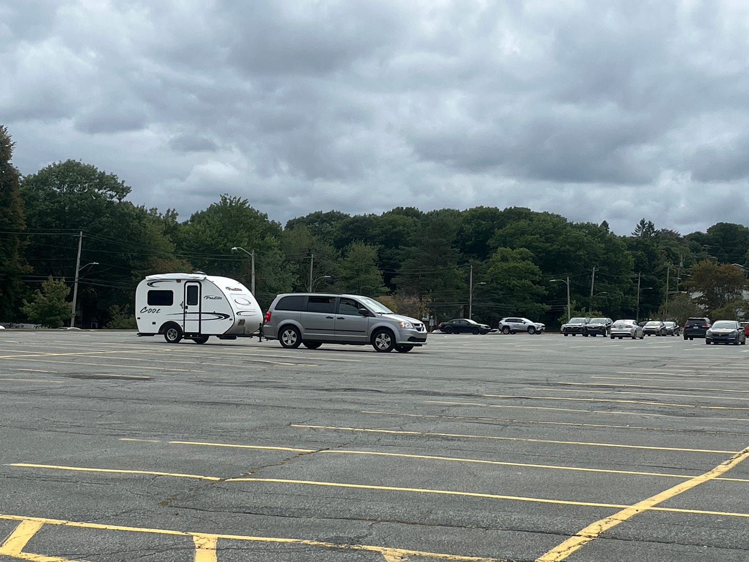 Our grey Dodge Grand Caravan parked in a mostly empty parking lot with a white little camper trailer behind it. It is as cute as a button.