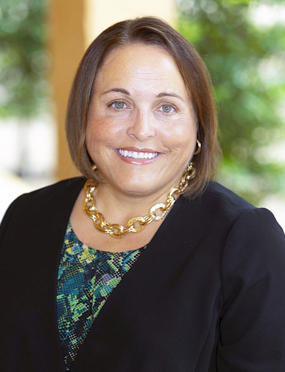 A head-and-shoulders professional portrait shows a person facing the camera and smiling directly at the viewer. They have shoulder-length brown hair, wear a black blazer over a blue and green patterned top, and a large gold chain necklace with matching hoop earrings. The background is softly blurred with green foliage and warm architectural elements, drawing attention to the subject's face.