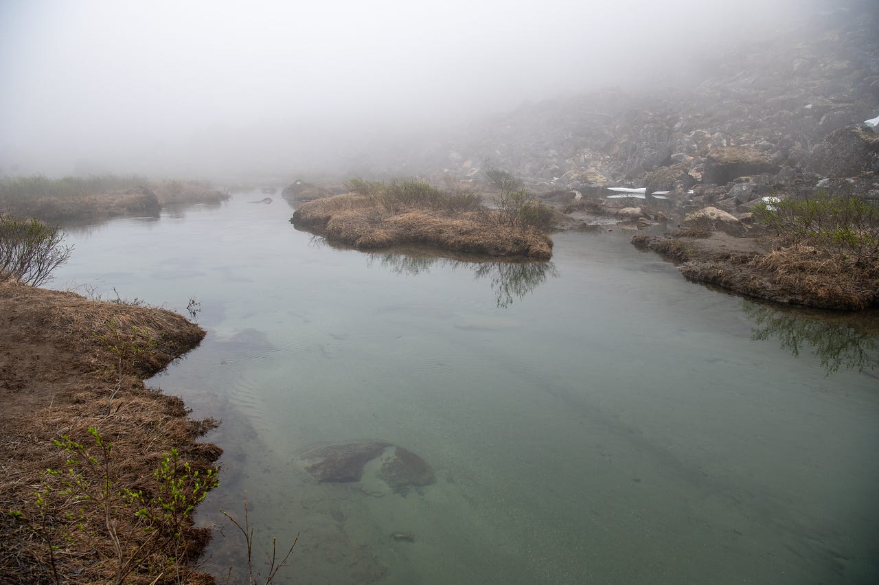 A calm, clear stream winds between low banks of brown grass and tundra, its water a pale glassy green over sunken boulders. Sparse green leaves are just emerging on the bank brush. Upstream, patches of ice cling among rocks at the base of a talus slope that fades upward into thick fog.