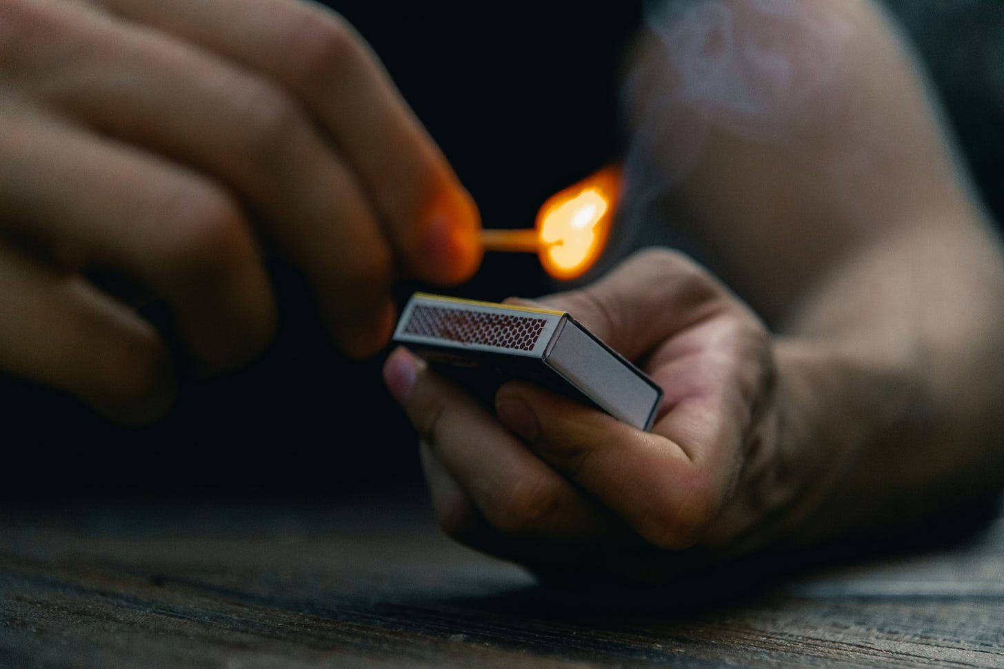 Two hands holding a matchbox and one match is being lit and has a flame.