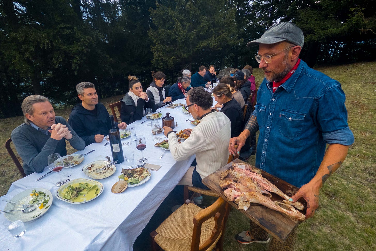 Eine lange Tafel ist im Freien auf einer Wiese am Waldrand gedeckt, 25 Personen sitzen eng nebeneinander und geniessen ein gemeinsames Abendessen. Im Vordergrund rechts steht Spitzenkoch Simon Schneeberger mit Jeanshemd und tätowierten Armen, der ein Holzbrett mit Gitzi-Gigot serviert. Auf dem Tisch stehen Weingläser, leere und halbvolle Teller sowie Karaffen. Die Stimmung wirkt lebendig, gesellig und rustikal – ein gemeinsames Essen in der Natur. Eine lange Tafel ist im Freien auf einer Wiese am Waldrand gedeckt, 25 Personen sitzen eng nebeneinander und geniessen ein gemeinsames Abendessen. Im Vordergrund rechts steht Spitzenkoch Simon Schneeberger mit Jeanshemd und tätowierten Armen, der ein Holzbrett mit Gitzi-Gigot serviert. Auf dem Tisch stehen Weingläser, leere und halbvolle Teller sowie Karaffen. Die Stimmung wirkt lebendig, gesellig und rustikal – ein gemeinsames Essen in der Natur.
