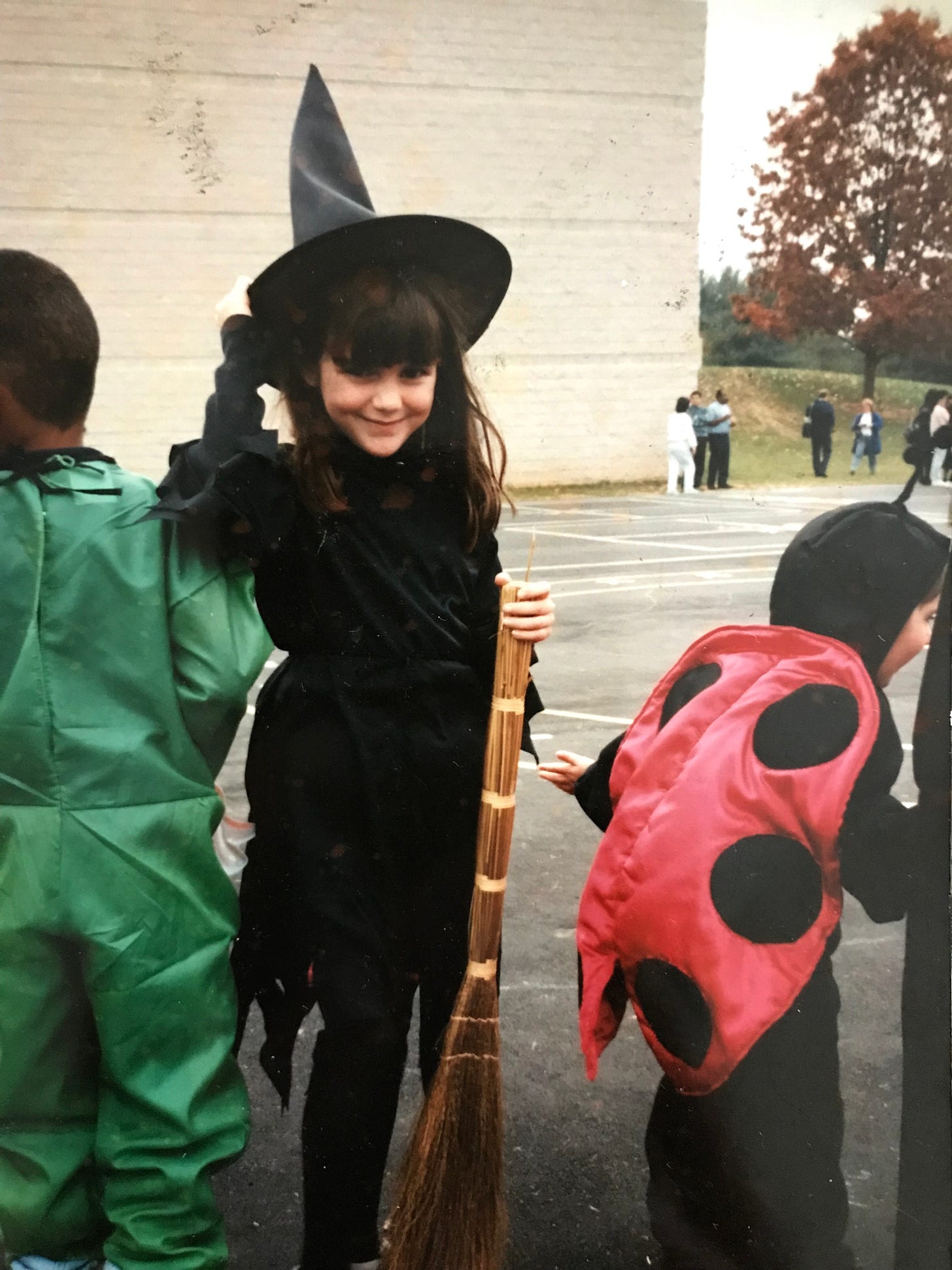 An image of little girl molly standing in a halloween parade in a witch costume, smiling and holding one hand on her witches hat and the other holding a straw broom