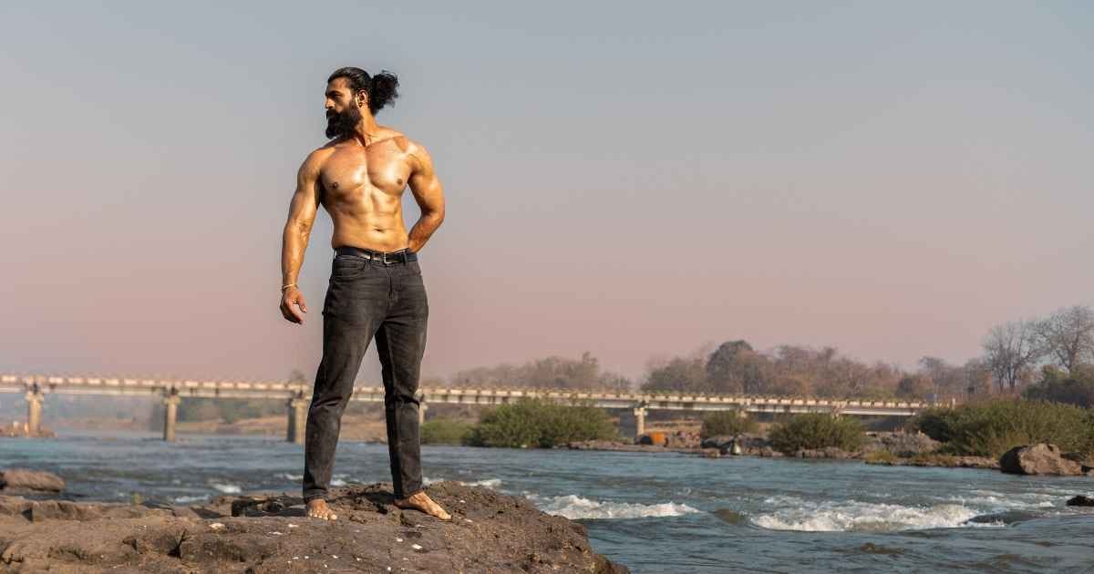 A shirtless muscular man standing confidently on rocks beside a serene river with a bridge in the background A shirtless muscular man standing confidently on rocks beside a serene river with a bridge in the background