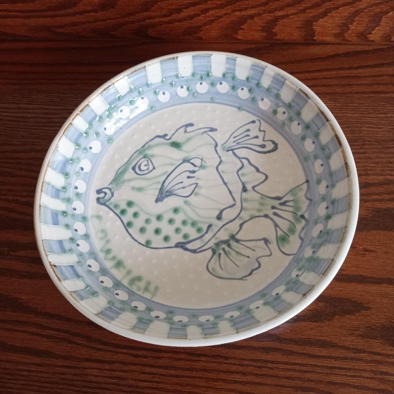 May include: A ceramic plate with a hand-painted fish design. The fish is depicted in shades of green and blue, with a whimsical style. The plate has a blue and white checkered border and a dotted pattern. The word "WEIGH" is written on the plate.